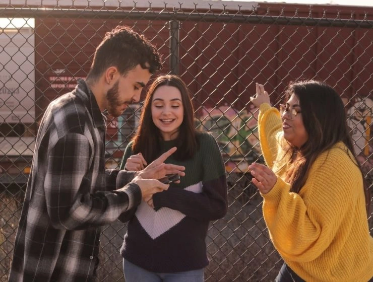 Tres jóvenes charlando y gesticulando delante de una valla metálica