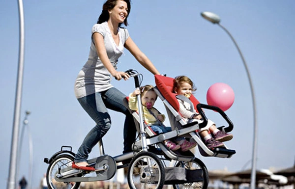 Madre sonriendo mientras pasea a sus dos hijos en una innovadora carriola bicicleta, un invento para padres activos que combina ejercicio y diversión.