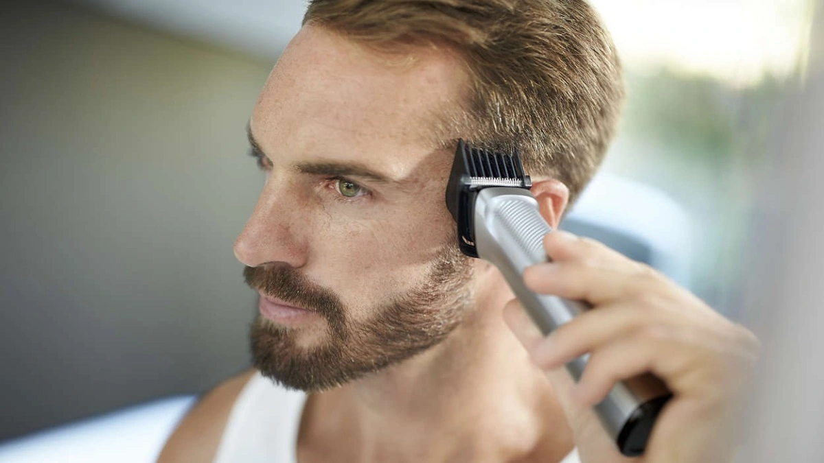 Mantenimiento del Corte de Pelo: Consejos para Cuidar tu Mullet Hombre recortando el cabello de la patilla y el contorno de la oreja con una máquina de cortar pelo eléctrica, mostrando el cuidado personal y el mantenimiento de un peinado masculino.