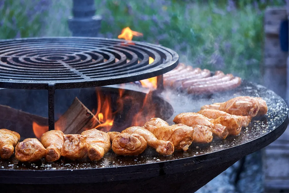 El centro de la reunión: una parrilla para cocinar y socializar. Asador redondo tipo plancha con fuego de leña central, cocinando muslos de pollo y chorizos en sus diferentes zonas de calor.