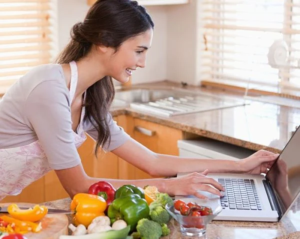 Planificación Digital de Comidas Saludables Mujer planificando su menú semanal en la laptop, con vegetales frescos en la encimera listos para cocinar.
