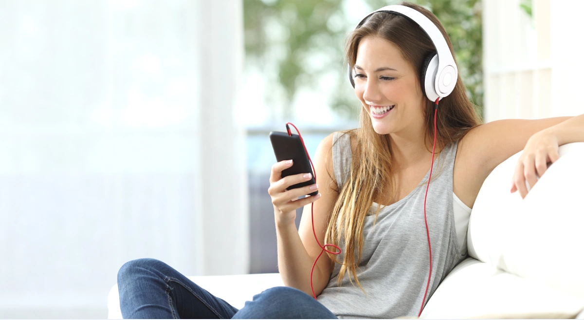 Mujer escuchando música con auriculares blancos conectados a su teléfono mientras sonríe relajada en el sofá.