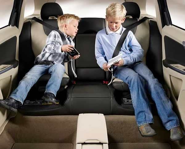 Dos niños rubios sentados en el asiento trasero de un coche con cinturón, jugando con aparatos