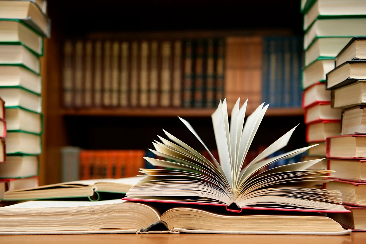 Libros apilados y un libro abierto en abanico sobre una mesa, con estantería de madera detrás