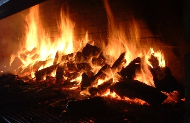 El secreto del auténtico sabor a la parrilla: el fuego de leña. Primer plano de leña ardiendo con llamas intensas dentro de una parrilla de leña, creando brasas para un asado tradicional.