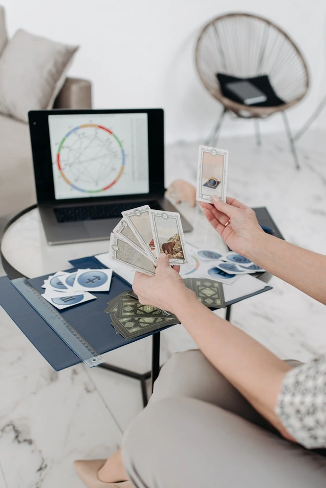 Manos sosteniendo cartas de tarot frente a un portátil que muestra una carta astral