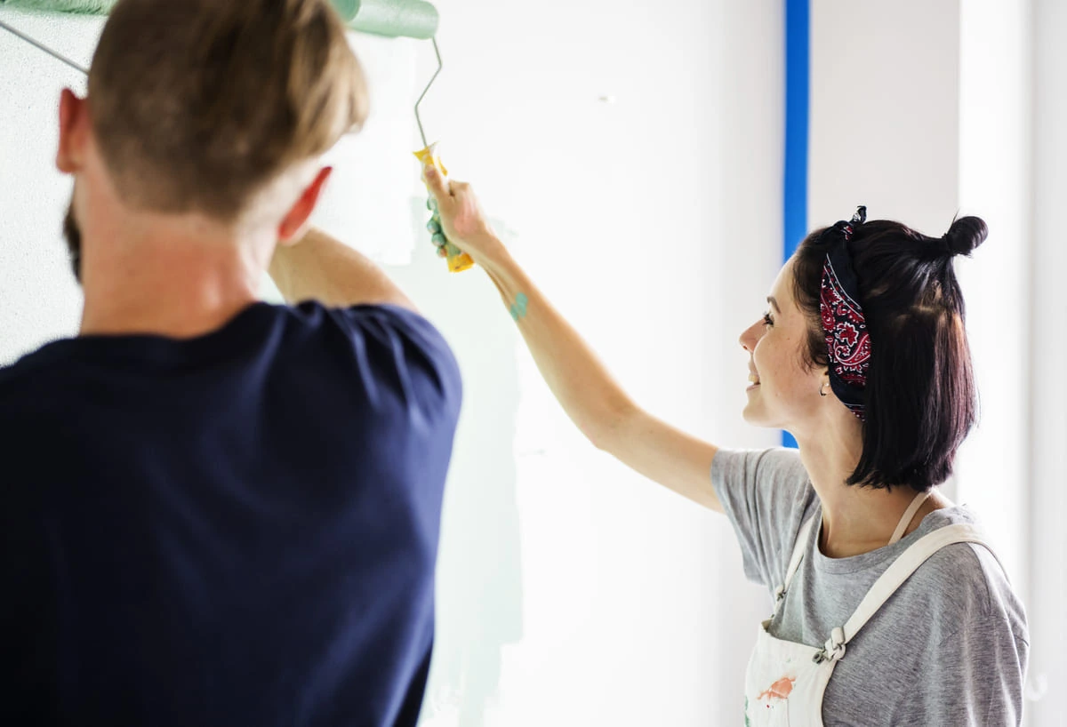 Prueba el Color Antes de Decidir Pareja sonriendo mientras prueba una muestra de pintura en la pared para elegir el color correcto y evitar errores de decoración.