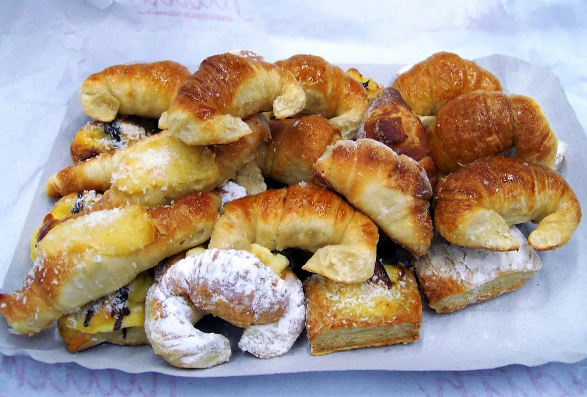 Una charola con papel blanco llena de un surtido de pan dulce, incluyendo medialunas de manteca glaseadas, facturas con crema pastelera y cuernitos con azúcar glass.