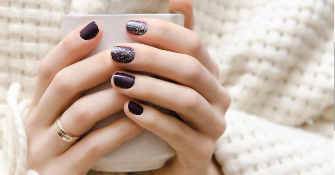 Diseño de Uñas Elegantes para Otoño. Manos de mujer con un elegante diseño de uñas en color berenjena o morado oscuro, con una uña de acento con glitter, sosteniendo una taza sobre un suéter de punto.