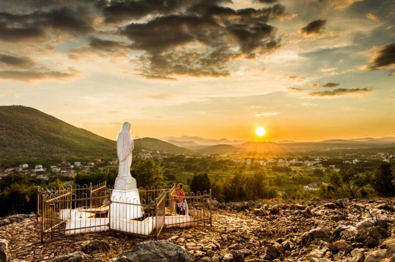 Virgen María en colina con amanecer y vista panorámica del valle.

