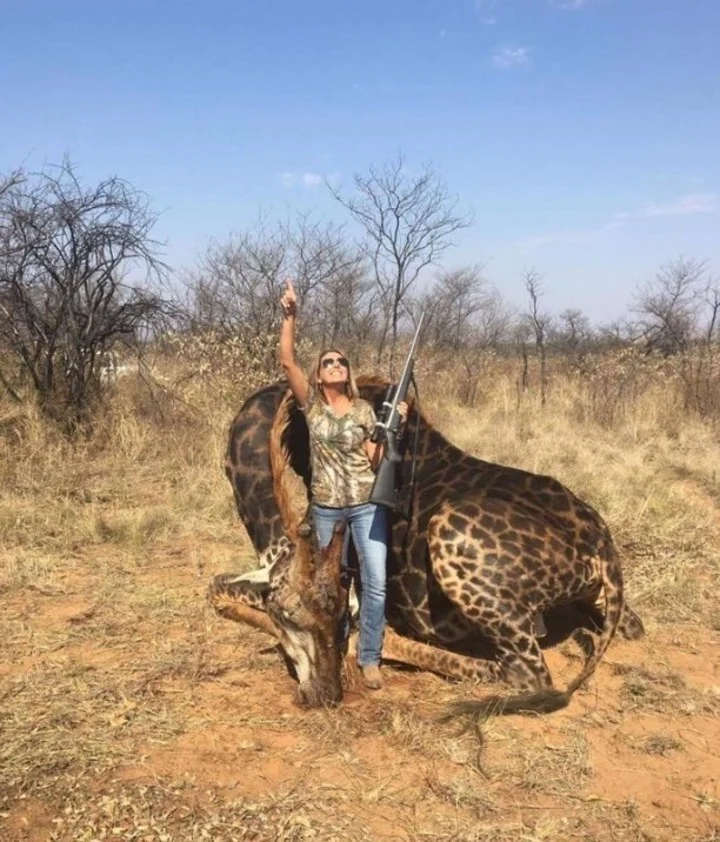 Mujer con rifle posa señalando al cielo junto a una jirafa abatida en la sabana
