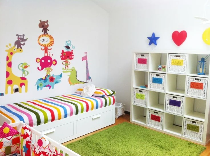 Habitación infantil colorida y organizada con vinilos de animales en la pared, una estantería con cestas etiquetadas para juguetes y una cama con cajones.