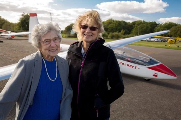 Mujer mayor sonriente junto a una piloto, delante de un planeador en un aeródromo