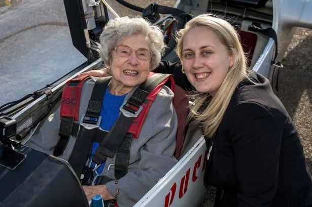 Anciana sonriente con arnés rojo en la cabina de un planeador, junto a una joven