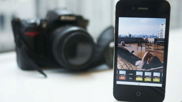 Las Mejores Apps para Editar Fotos en 2026 Primer plano de un smartphone usando una app para editar fotos y aplicar filtros, con una cámara réflex profesional desenfocada en el fondo.